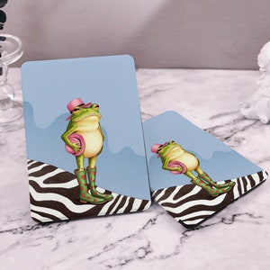 May include: Two blue and white striped coasters with a green frog wearing a pink hat and holding a pink umbrella. The frog is wearing red boots with white hearts.