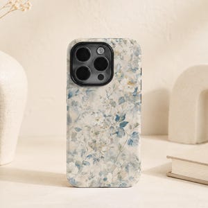 Könnte beinhalten: Eine Smartphone-Hülle mit einem weichen, aquarellartigen Blumenmuster in gedeckten Blau-, Creme- und Weißtönen. Das Design bedeckt die gesamte Rückseite der Hülle, die oben links einen schwarzen Ausschnitt für die Kamera aufweist.