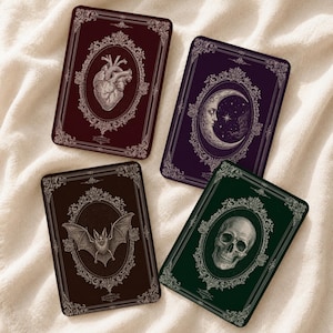 Puede incluir: Cuatro tarjetas rectangulares con marcos ornamentados complejos. Una tarjeta muestra un corazón anatómico sobre fondo rojo oscuro, una una luna creciente sobre morado, una un murciélago sobre marrón oscuro y una un cráneo humano sobre fondo verde oscuro.
