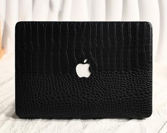May include: A black laptop cover with a textured crocodile skin pattern on the top half and a pebbled leather texture on the bottom half, featuring a central cutout that reveals the white Apple logo on the laptop underneath the protective case.