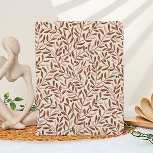 May include: A rectangular notebook with a cream-colored background and a repeating pattern of brown leaves and green olives. The notebook is displayed on a white surface with a decorative statue and an open book in the background.