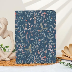 Puede incluir: Un cuaderno con un estampado floral azul oscuro con flores y hojas rosas, verdes y blancas. El diseño es un patrón repetitivo de elementos botánicos. El cuaderno es rectangular y parece estar cerrado.