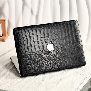 May include: Black laptop case with a crocodile skin texture and a white Apple logo. The case is on a white surface with a calendar and a cup of coffee in the background. The case is designed to protect the laptop.