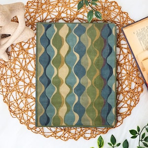 May include: A rectangular rug with a wavy pattern in shades of blue, green, and beige. The rug is placed on a woven placemat, with a book and decorative elements nearby. The rug's design features vertical, flowing lines.