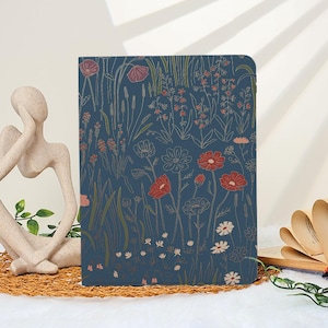 May include: A navy blue journal with a floral design featuring red, white, and gold flowers and foliage. The journal is closed and stands upright on a woven mat. A decorative sculpture and an open book are in the background.