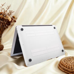 May include: A white laptop computer with a black screen is propped open, revealing the underside of the device. The bottom of the laptop has ventilation slots and four black rubber feet. The background is a cream-colored fabric.