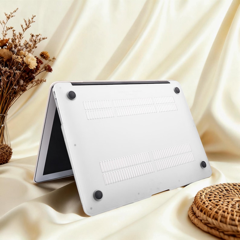 May include: A white laptop computer with a black screen is propped open, revealing the underside of the device. The bottom of the laptop has ventilation slots and four black rubber feet. The background is a cream-colored fabric.