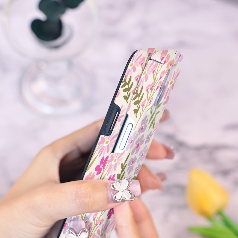 Puede incluir: Una funda de tel&eacute;fono floral con un estampado de flores rosas y moradas y hojas verdes sobre un fondo crema. La funda tiene un borde negro y se sostiene en una mano.