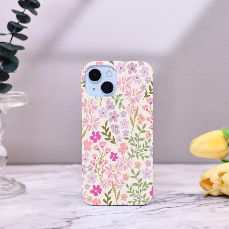 Puede incluir: Funda de tel&eacute;fono con dise&ntilde;o floral. El dise&ntilde;o presenta flores rosas, moradas y melocot&oacute;n con hojas verdes sobre un fondo crema. La funda del tel&eacute;fono tiene un m&oacute;dulo de c&aacute;mara azul claro.