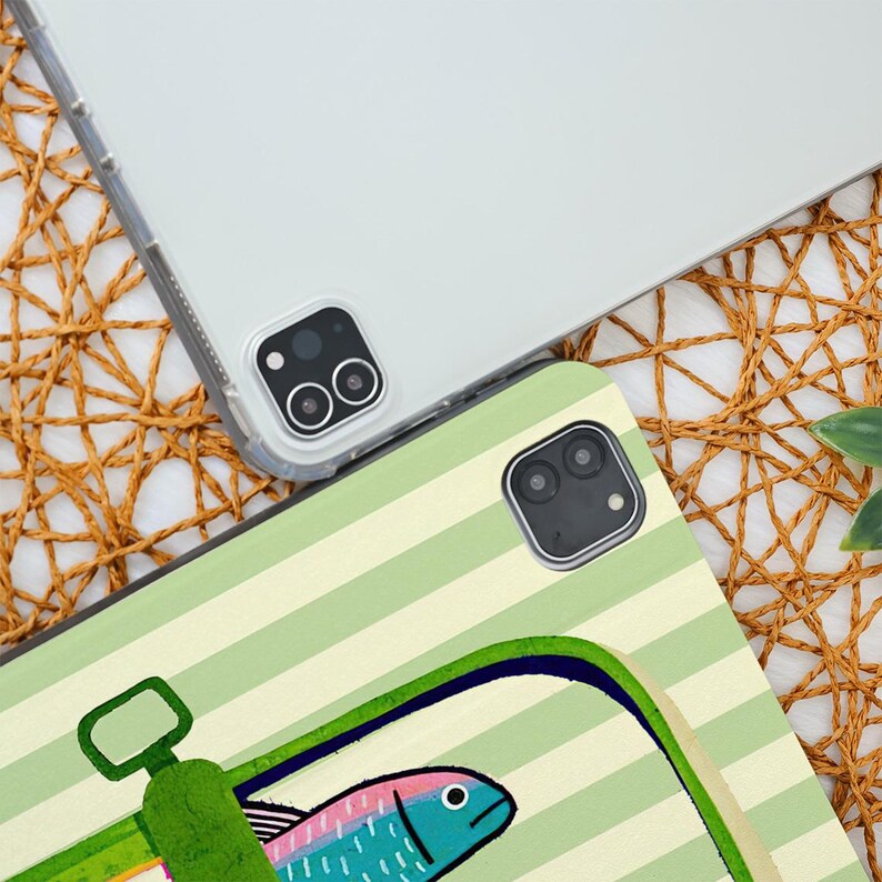 May include: Two tablet cases. One is clear, the other has a green and white striped pattern with a sardine can illustration. The sardine has blue, pink, and yellow details. The cases are on a woven surface.
