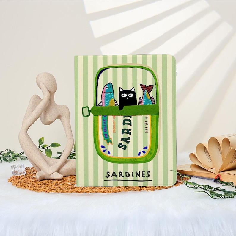 May include: A notebook with a sardine can design. The cover features a green and white striped background with a sardine can illustration. The can contains two colorful fish and a black cat. The word "Sardines" is printed on the cover.