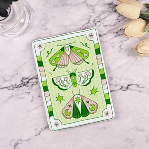 May include: A green and pink card with a white border features three stylized butterflies. The butterflies have green wings with pink accents and are surrounded by stars, moons, and mushrooms.