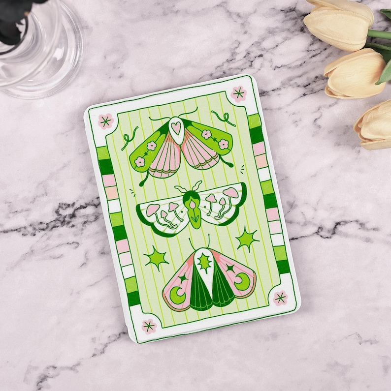 May include: A green and pink card with a white border features three stylized butterflies. The butterflies have green wings with pink accents and are surrounded by stars, moons, and mushrooms.