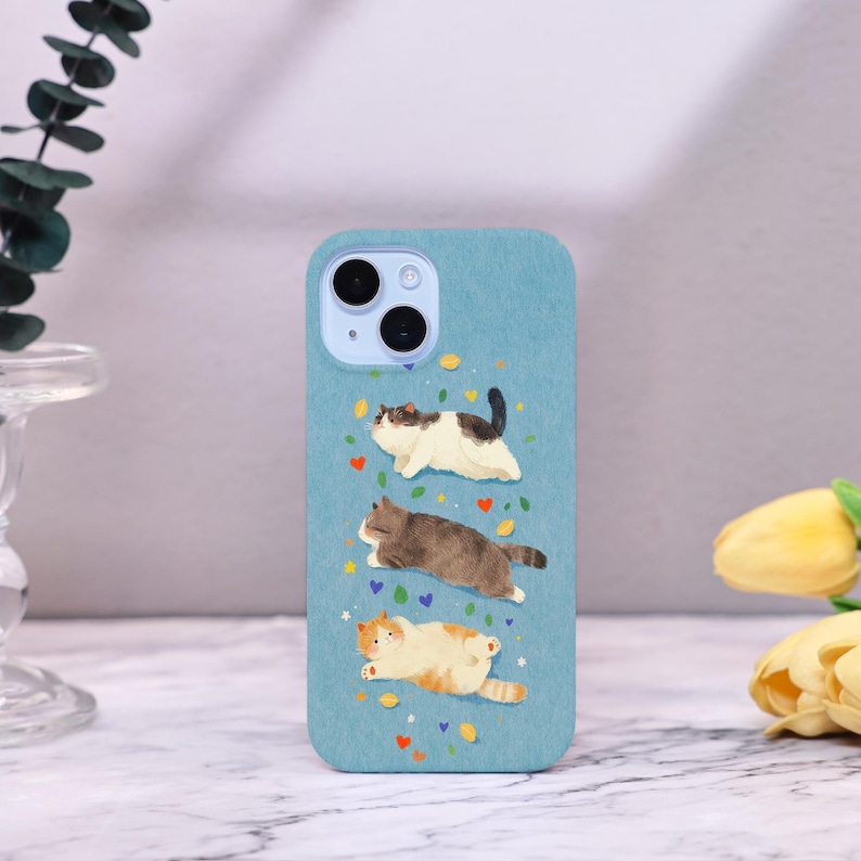 Op de afbeelding: Lichtblau telefoonhoesje met een grillige illustratie van drie katten in verschillende kleuren. De katten worden afgebeeld in een ontspannen pose, met kleurrijke harten en sterren. De hoes heeft een uitsparing voor een dubbele camera.