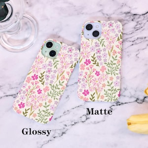 Puede incluir: Dos fundas para tel&eacute;fono con un estampado floral en rosa, morado y verde sobre un fondo crema. Una funda est&aacute; etiquetada como "Glossy" y la otra como "Matte". Las fundas est&aacute;n sobre una superficie de m&aacute;rmol.