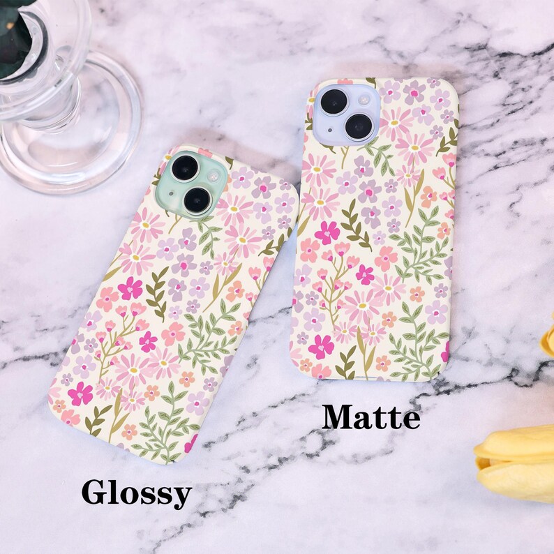 Puede incluir: Dos fundas para tel&eacute;fono con un estampado floral en rosa, morado y verde sobre un fondo crema. Una funda est&aacute; etiquetada como "Glossy" y la otra como "Matte". Las fundas est&aacute;n sobre una superficie de m&aacute;rmol.
