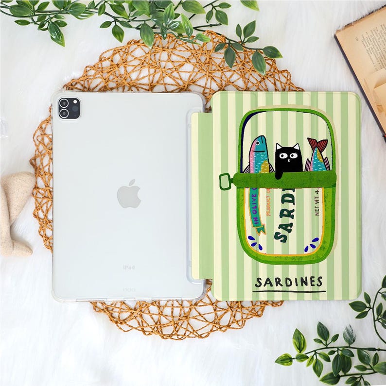 May include: A clear iPad case with a sardine can design. The sardine can illustration features a black cat and two colorful fish inside a green can with the word "Sardines" printed on it. The case is on a white surface.