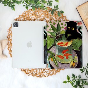 May include: A white iPad with a clear case next to a tablet case featuring a painting of a black cat with yellow eyes, looking at goldfish in a glass bowl. The case has a floral and leaf design.