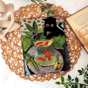 May include: A decorative art print featuring a black cat with yellow eyes perched above a bowl of goldfish. The artwork has a colorful, painterly style with green foliage and a floral background. The print is displayed on a woven placemat.