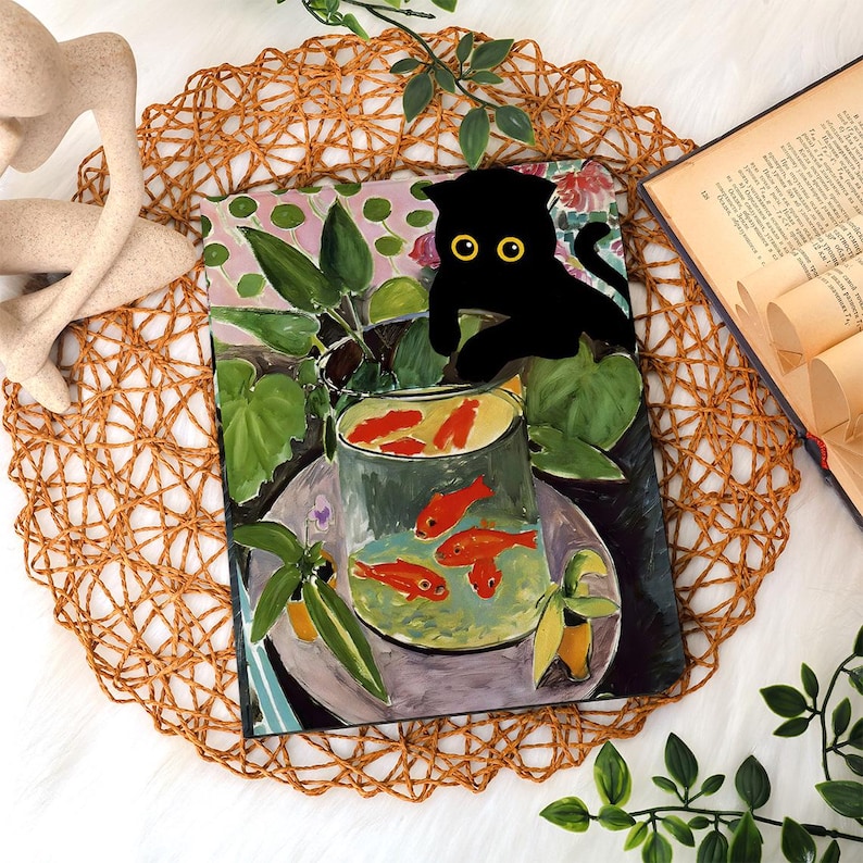 May include: A decorative art print featuring a black cat with yellow eyes perched above a bowl of goldfish. The artwork has a colorful, painterly style with green foliage and a floral background. The print is displayed on a woven placemat.