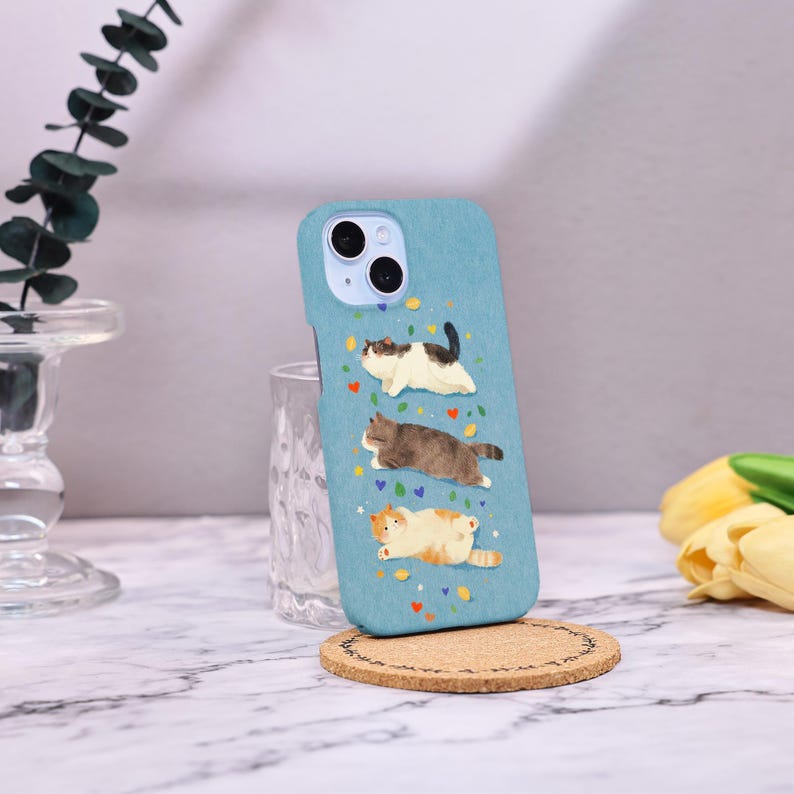Op de afbeelding: Lichtblauwe telefoonhoes met drie cartoonkatten in verschillende poses. De katten zijn wit, bruin en oranje, met kleurrijke hart- en bladaccenten. De hoes ligt op een ronde kurken onderzetter.