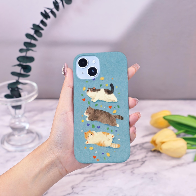 Op de afbeelding: Lichtblauwe telefoonhoes met drie katten in aquarel. De katten zijn wit, bruin en oranje, met kleurrijke hart- en bladaccenten. De hoes heeft een uitsparing voor een camera met dubbele lens.
