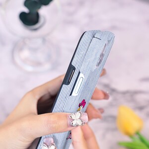 Może przedstawiać: Niebiesko-białe etui na telefon w paski z postacią z kresk&oacute;wki i r&oacute;żową kokardą. Etui ma czarną obw&oacute;dkę i srebrny guzik. Etui jest trzymane przez osobę.