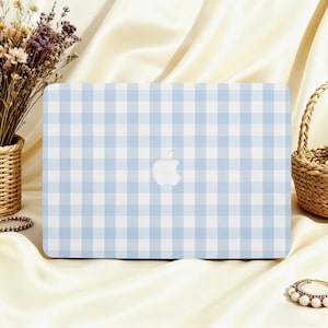 Peut inclure: Coque d'ordinateur portable à motif vichy bleu clair et blanc, avec le logo Apple au centre. La coque est posée sur une surface en satin crème, avec un panier en osier et des fleurs séchées en arrière-plan.