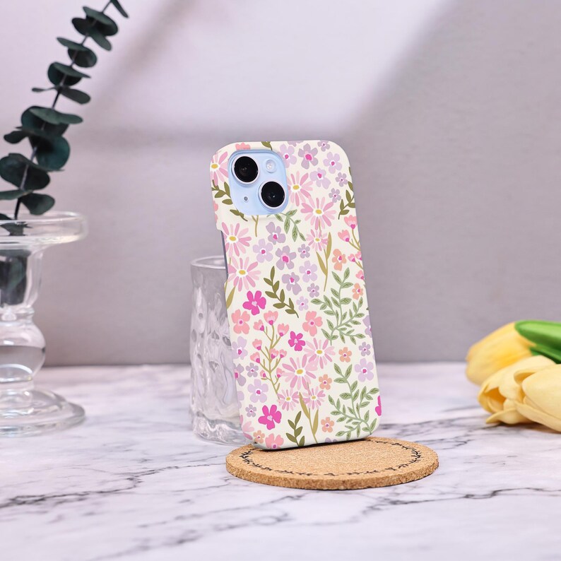 Puede incluir: Una funda de tel&eacute;fono floral con un patr&oacute;n de flores rosas, moradas y blancas con hojas verdes. La funda est&aacute; sobre un posavasos de corcho, con un vaso transparente y una superficie de m&aacute;rmol en el fondo.