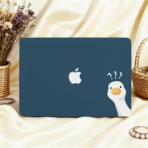 May include: A navy blue laptop case featuring a white Apple logo and a cartoon goose with question marks. The goose has a yellow beak and is peeking from the side. The case is on a cream-colored surface.