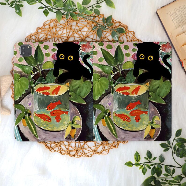 May include: A tablet case featuring a whimsical design. The case showcases a black cat silhouette with yellow eyes, perched above a goldfish bowl. The background includes a floral and foliage pattern with a pink and green color scheme.