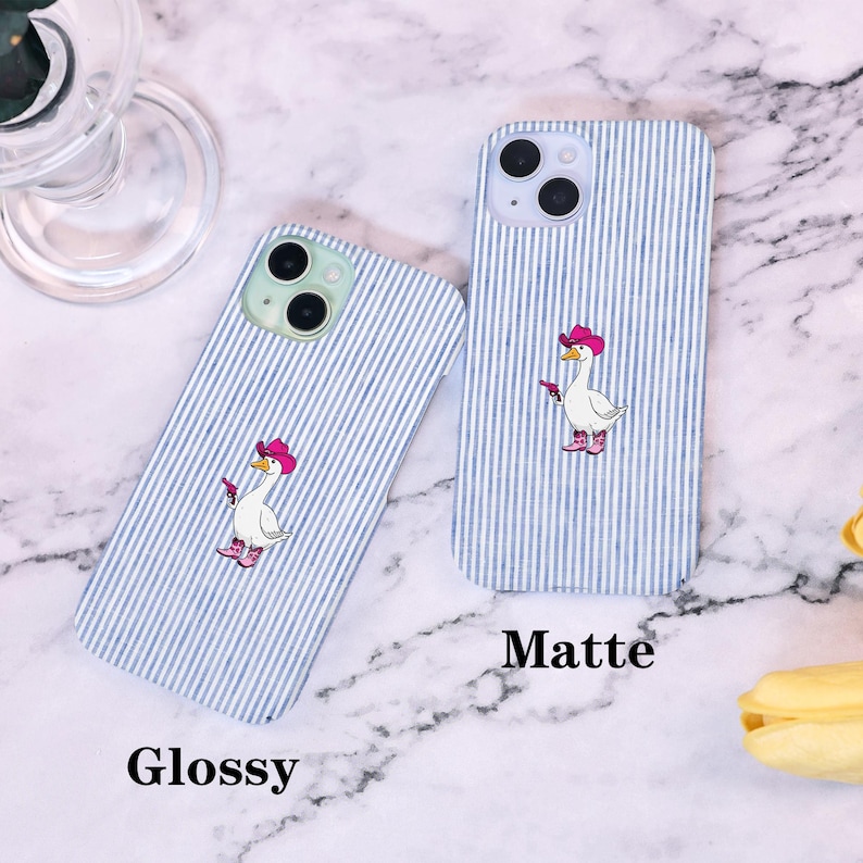 Może przedstawiać: Dwa etui na telefon z niebiesko-białym wzorem w paski. Jedno etui jest oznaczone jako "Glossy", a drugie jako "Matte". Każde etui przedstawia kresk&oacute;wkową gęś w r&oacute;żowym kapeluszu kowbojskim i butach, trzymającą pistolet.