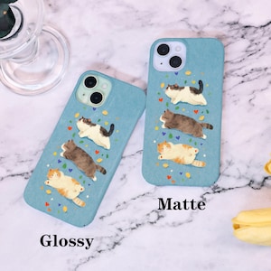 Op de afbeelding: Twee blauwe telefoonhoesjes met een kattenontwerp. E&eacute;n hoesje is gemarkeerd met "Glossy" en de andere met "Matte". Elk hoesje heeft drie cartoonkatten in verschillende kleuren, met kleurrijke confetti en hartaccenten.
