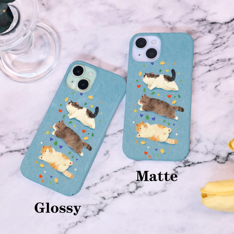 Op de afbeelding: Twee blauwe telefoonhoesjes met een kattenontwerp. E&eacute;n hoesje is gemarkeerd met "Glossy" en de andere met "Matte". Elk hoesje heeft drie cartoonkatten in verschillende kleuren, met kleurrijke confetti en hartaccenten.