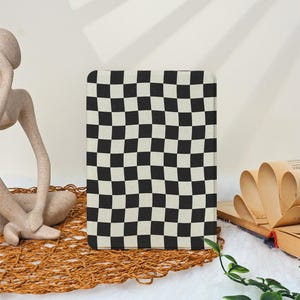 May include: A rectangular notebook featuring a black and white chequerboard design. The notebook is positioned upright on a woven mat, with a sculpture and an open book in the background.
