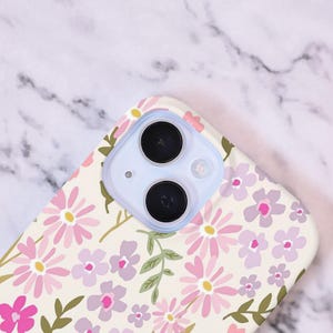 Puede incluir: Funda de tel&eacute;fono floral con fondo crema, adornada con flores rosas y moradas y hojas verdes. La funda tiene un recorte para la lente de la c&aacute;mara con un borde azul claro.