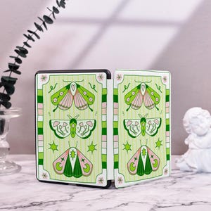 May include: A green and pink book cover with a butterfly design. The cover is divided into two sections, each with a different butterfly pattern. The butterflies are surrounded by green and pink flowers and leaves.