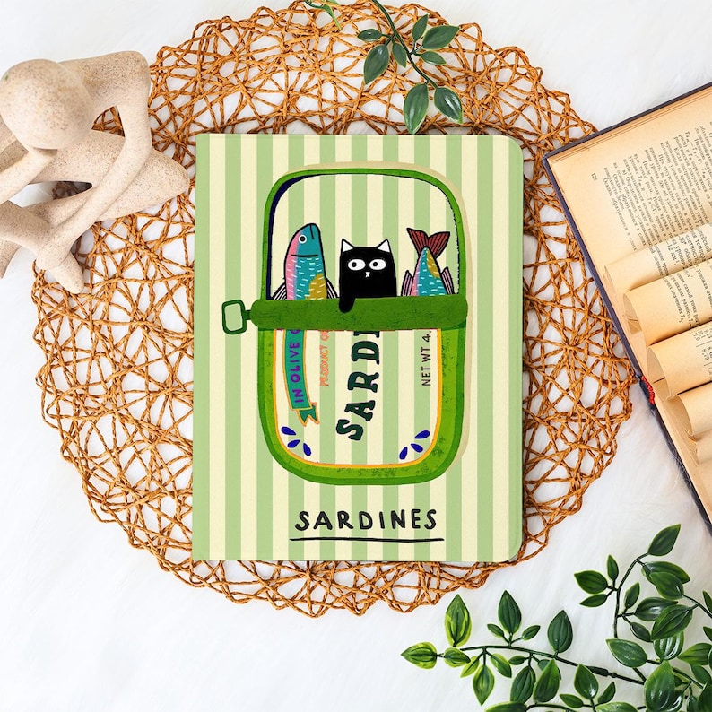 May include: Notebook with a whimsical sardine can design. The illustration features a black cat peeking out between two colorful fish inside a green can, set against a striped background. The word "Sardines" is printed below.