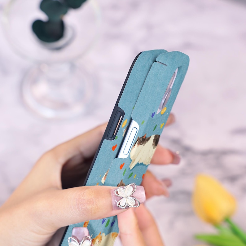 Op de afbeelding: Lichtblauwe telefoonhoes met een kattenillustratie. De hoes heeft een zwarte rand en een uitsparing voor de camera. Het ontwerp bevat kleurrijke confetti-vormen. De hoes wordt door een persoon vastgehouden.