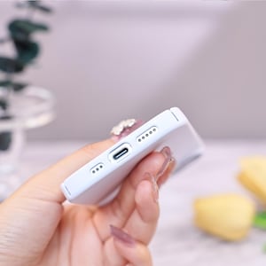 Puede incluir: Una funda de tel&eacute;fono azul claro con un puerto de carga y orificios para altavoces. La funda se sostiene en una mano, mostrando su dise&ntilde;o elegante y superficie lisa. La funda est&aacute; dise&ntilde;ada para proteger el tel&eacute;fono de ara&ntilde;azos y da&ntilde;os.