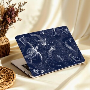 May include: A navy blue laptop skin featuring a white illustration of various marine animals, including a sea turtle, jellyfish, and whale. The design includes a starry background. The laptop is open on a beige surface.