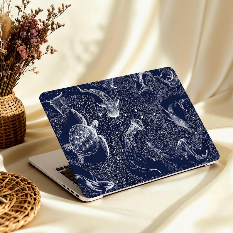 May include: A navy blue laptop skin featuring a white illustration of various marine animals, including a sea turtle, jellyfish, and whale. The design includes a starry background. The laptop is open on a beige surface.