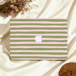 May include: A laptop case with a design of olive green and light pink horizontal stripes. The Apple logo is white. The case is on a white satin surface, with dried flowers and a woven coaster in the background.