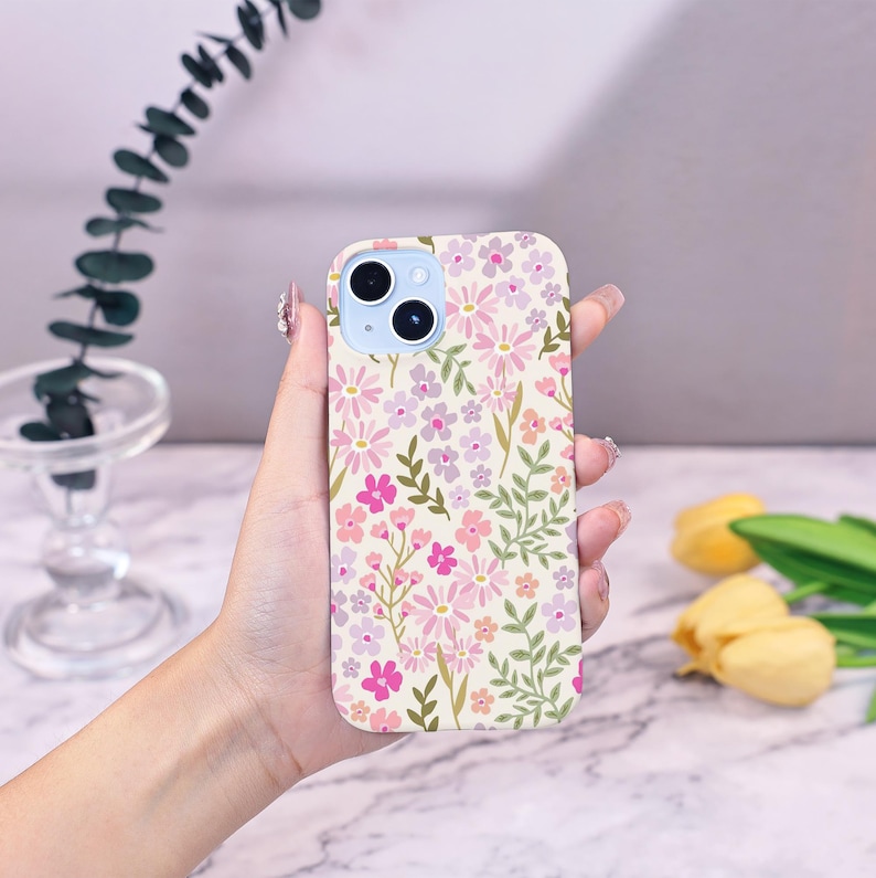 Puede incluir: Una funda floral para tel&eacute;fono con un estampado de flores rosas, moradas y blancas y hojas verdes sobre un fondo crema. La funda tiene un m&oacute;dulo de c&aacute;mara azul claro y se sostiene en una mano.