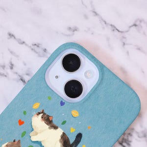 Op de afbeelding: Een lichtblauwe telefoonhoes met een grillige illustratie van een kat die omhoog kijkt. De kat is wit en bruin, met kleurrijke confetti-achtige vormen. De hoes heeft een camera-uitsparing.