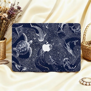 Peut inclure: Une coque d'ordinateur portable bleu marine avec une illustration blanche d'animaux marins, dont une tortue, un requin-baleine et une méduse, sur un fond étoilé. Le logo Apple est visible. La coque est posée sur une surface crème.