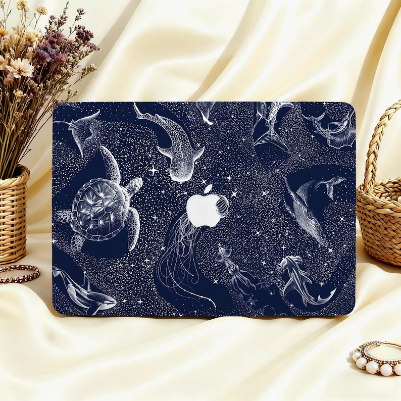 May include: A navy blue laptop case featuring a white illustration of various marine animals, including a turtle, whale shark, and jellyfish, set against a starry background. The Apple logo is visible. The case is on a cream-colored surface.