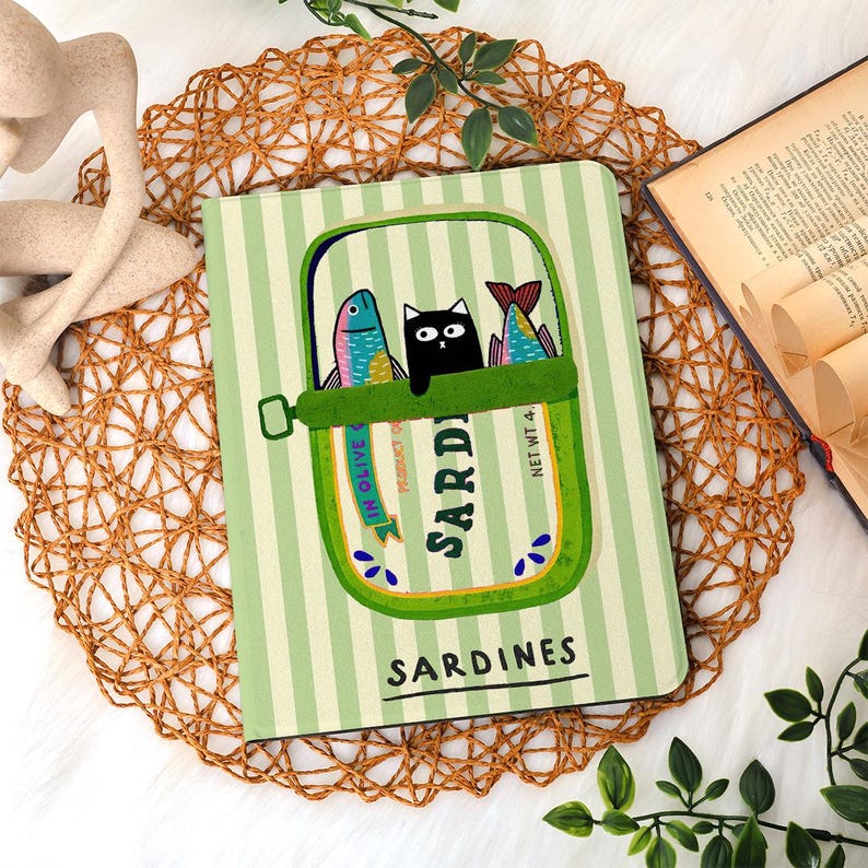 May include: A green and white striped notebook featuring a sardine can design with a black cat and two colorful fish. The words "Sardines" and "In Olive" are visible on the cover. The notebook is on a woven placemat.
