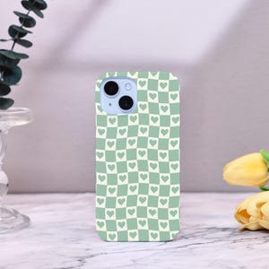 Könnte beinhalten: Eine Handyhülle mit einem grün-weißen Karomuster, wobei jedes Quadrat ein kleines Herz enthält. Die Hülle hat ein hellblaues Kameramodul und ist wahrscheinlich für ein Smartphone konzipiert. Das Gesamtdesign ist schlicht und niedlich.