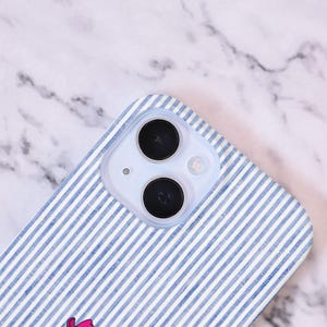 Może przedstawiać: Etui na smartfon z niebiesko-białym wzorem w paski. Etui posiada przezroczystą osłonę obiektywu aparatu z dwoma czarnymi obiektywami i małą lampą błyskową. Etui leży na białej i szarej marmurowej powierzchni.